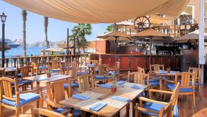 Un restaurante al aire libre con mesas y sillas de madera, una zona de bar y vistas al mar.