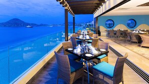 Un espacio de comedor al aire libre con sillas y mesas de mimbre, con vistas a una masa de agua y un paisaje urbano distante.
