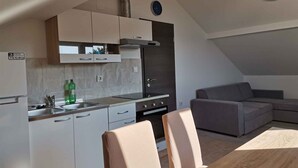 A compact kitchen with white cabinets, a stove, and a sink. There is a grey sofa and a wooden dining table with chairs.
