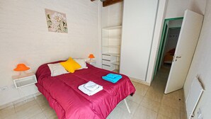 Un dormitorio con colcha roja, pared blanca con un cuadro floral, armario blanco y una puerta que da a otra habitación.