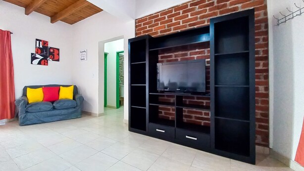 Un salón con televisión sobre una pared de ladrillo, sofá gris con cojines de colores y techo de madera.