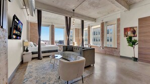A modern hotel room with a bed, sofa, and armchair, large windows with a city view, and a flat-screen TV on the wall. - Spacious Downtown Loft | Free Breakfast & Pool (Dallas)