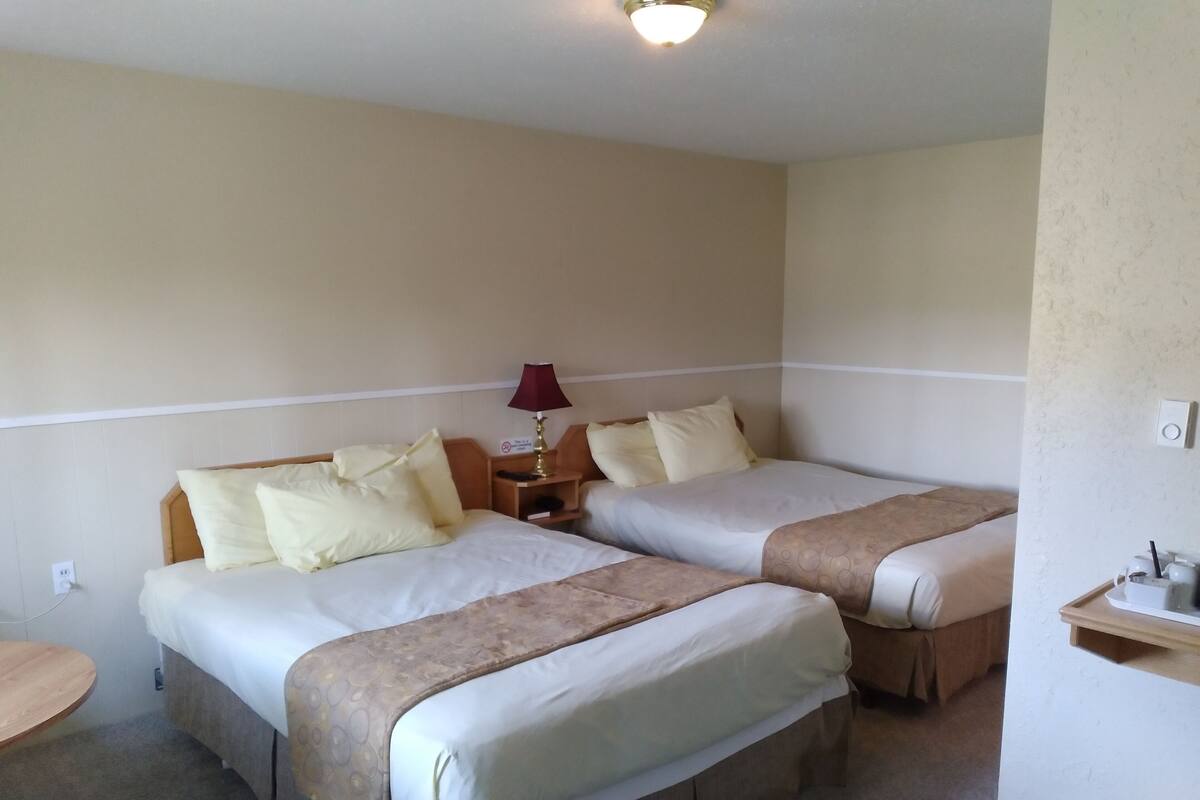 A hotel room with two beds, each with a headboard, a bedside table, a lamp, and a small shelf with toiletries.