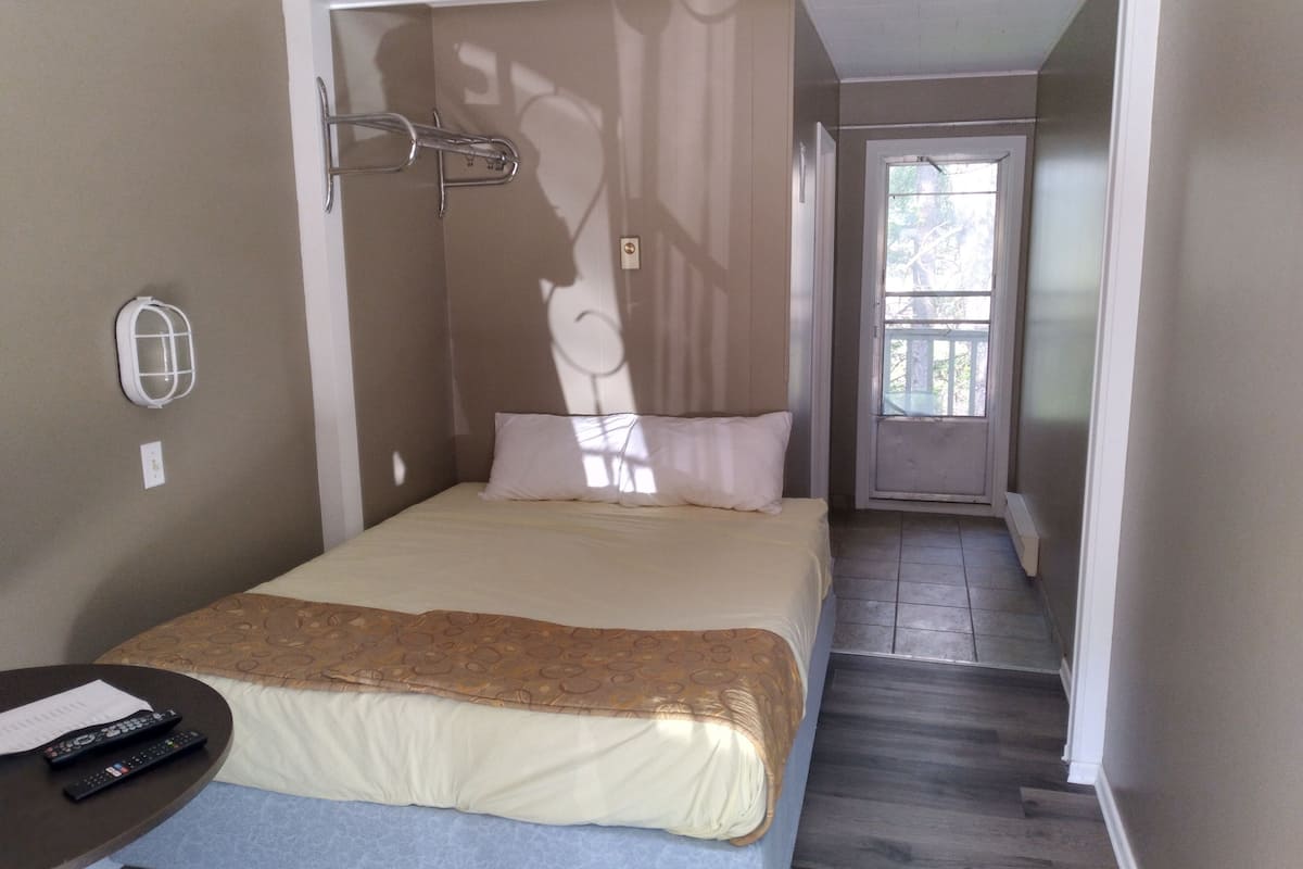A hotel room with a bed, a bedside table with a remote control, and a window with a view of the outside.
