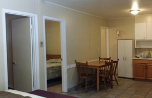 A compact room with a bed, a dining table with chairs, a kitchenette with cabinets, and a door leading to another room.