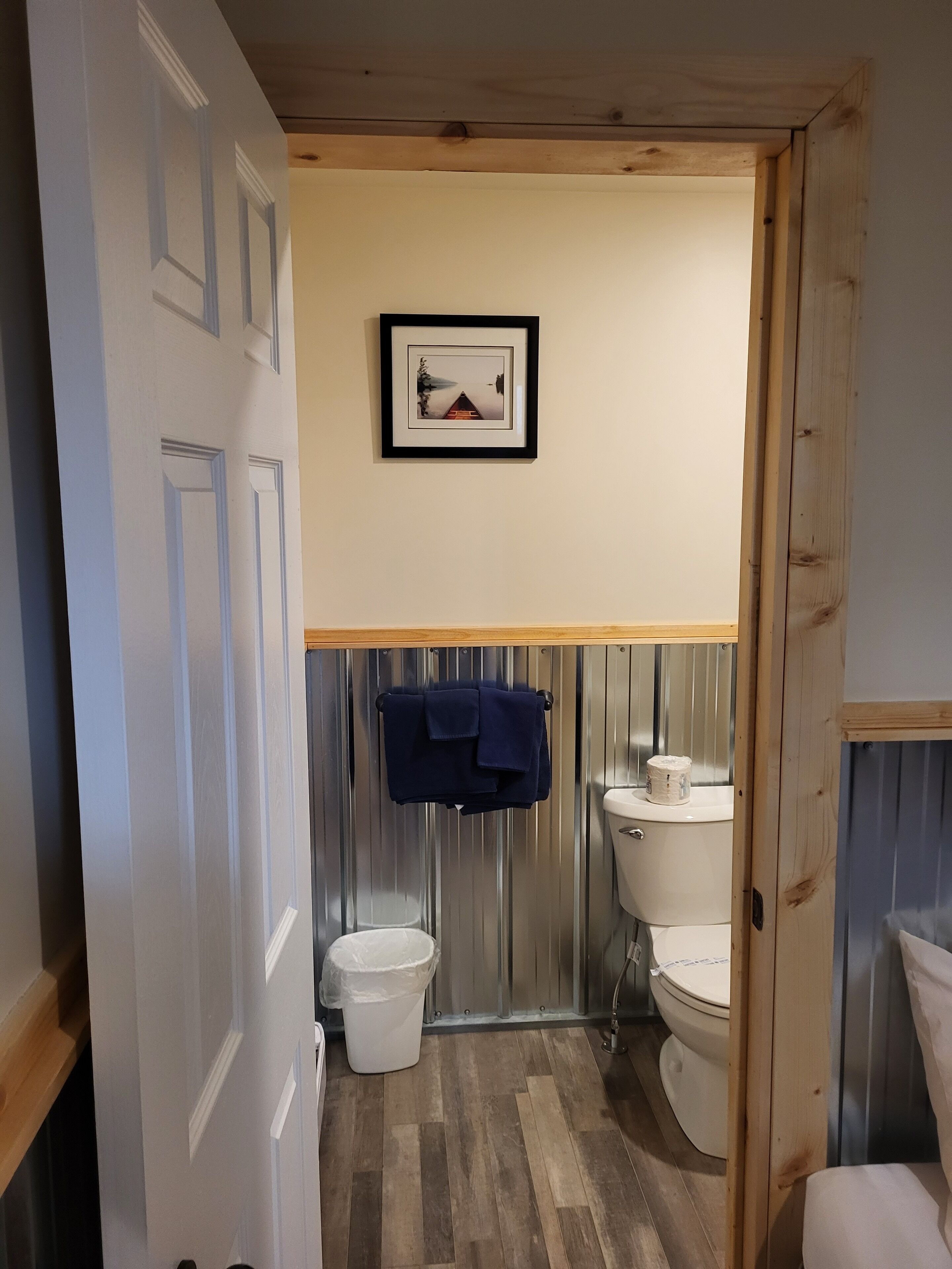 Un baño con techo de madera, un cuadro enmarcado en la pared, un inodoro blanco, un cubo blanco y una toalla azul colgada en un perchero.