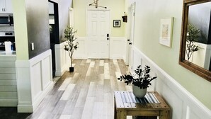 Un corridoio con pavimento in legno, pareti bianche e una parete verde decorativa. Si vede una porta in fondo al corridoio, un piccolo tavolo di legno con una pianta e uno specchio appeso al muro.