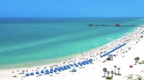 Una spiaggia con file di ombrelloni blu e lettini, un molo sullo sfondo e un cielo azzurro limpido.