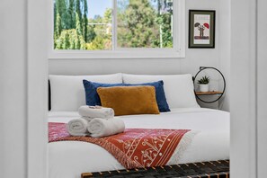 A neatly made bed with white linens, a brown throw blanket, and rolled towels. There is a window with a view of greenery outside.