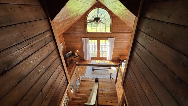 A wooden cabin with a loft bedroom, a staircase, a ceiling fan, and a window with curtains. - Cozy little cabin with creek access.  Great for fishing or tubing! (Landisburg)
