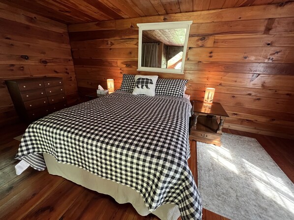 A wooden cabin bedroom with a checkered bedspread, a dresser, a nightstand with a lamp, and a mirror above the bed.