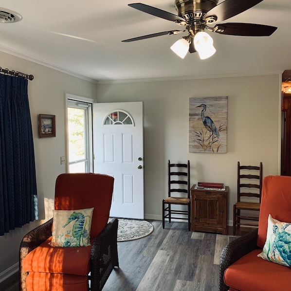 A living room with two wicker chairs, a ceiling fan, a white door, and a painting of a bird.