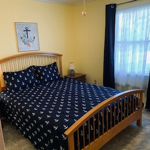 A bedroom with a wooden bed frame, navy blue bedding with anchor patterns, a bedside lamp, and a window with sheer curtains.