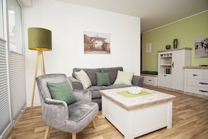 A modern living room with a grey sofa, a matching armchair, a white coffee table, and a green floor lamp. - Patio, free access to the AHOI adventure pool and sauna - Villa Karola, vacation apartment #02 - (Göhren)