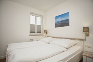 A double bed with white linens in a room with white walls, a window with blinds, a painting of a beach, and two wall-mounted lamps. - Patio, free access to the AHOI adventure pool and sauna - Villa Karola, vacation apartment #02 - (Göhren)