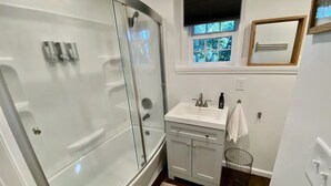 A bathroom with a glass-enclosed shower, a white sink with a mirror, and a small window with a view of trees. - Cute tiny home with everything you need! (Cedar Falls)