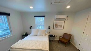 A bedroom with a bed, a chair, a small table, a window, an air conditioning unit, and a door. - Cute tiny home with everything you need! (Cedar Falls)