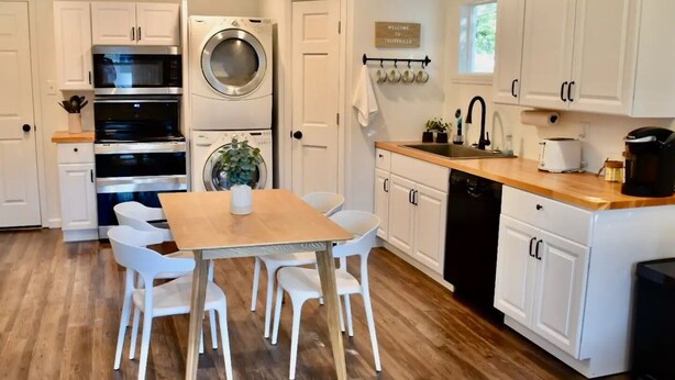 A modern kitchen with white cabinets, a wooden dining table, and a built-in oven. - Cute tiny home with everything you need! (Cedar Falls)