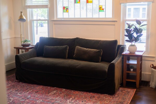 Ein dunkelgrünes Sofa mit zwei Kissen, ein gemustertes rotes Teppich, eine Stehlampe und ein kleiner Tisch mit einer Pflanze.