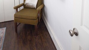Une pièce avec un plancher en bois, un fauteuil vert, une porte blanche, une plante suspendue et un miroir fixé au mur.