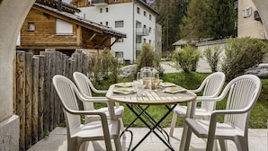 Une terrasse avec une table dressée pour quatre personnes, entourée de chaises en plastique blanches, d’où l’on peut admirer une montagne et un bâtiment en bois en arrière-plan.
