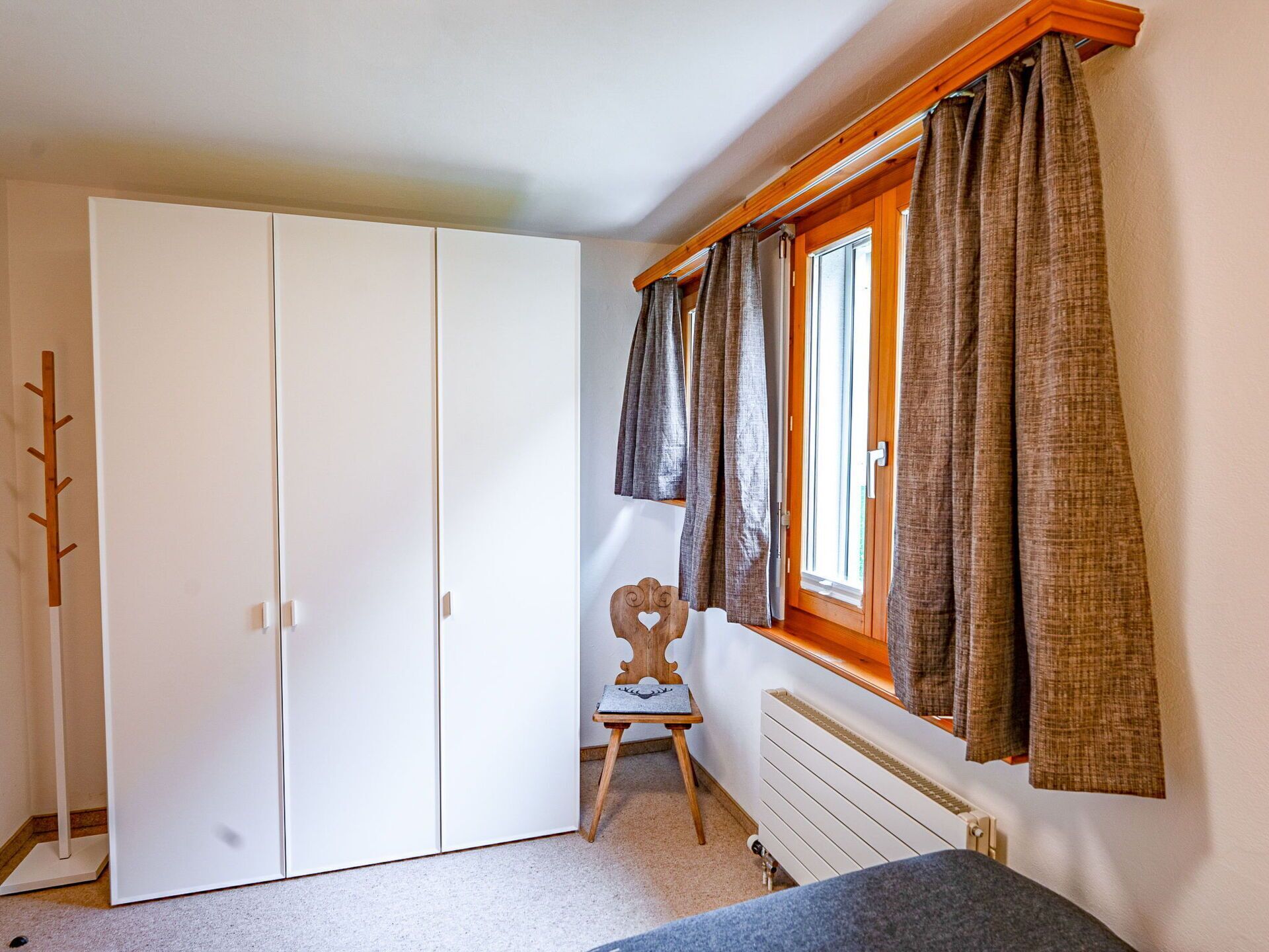 A bedroom with a large white wardrobe, a small wooden chair, a radiator, and a window with curtains.