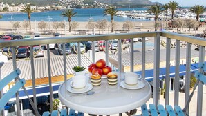 A balcony with a table set for breakfast, overlooking a marina and beach with parked cars and palm trees.