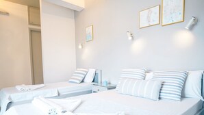 A hotel room with two beds, each with white bedding and striped pillows. There are two framed artworks on the wall and a bedside table with a lamp. - Rodi blue (Malevizi)