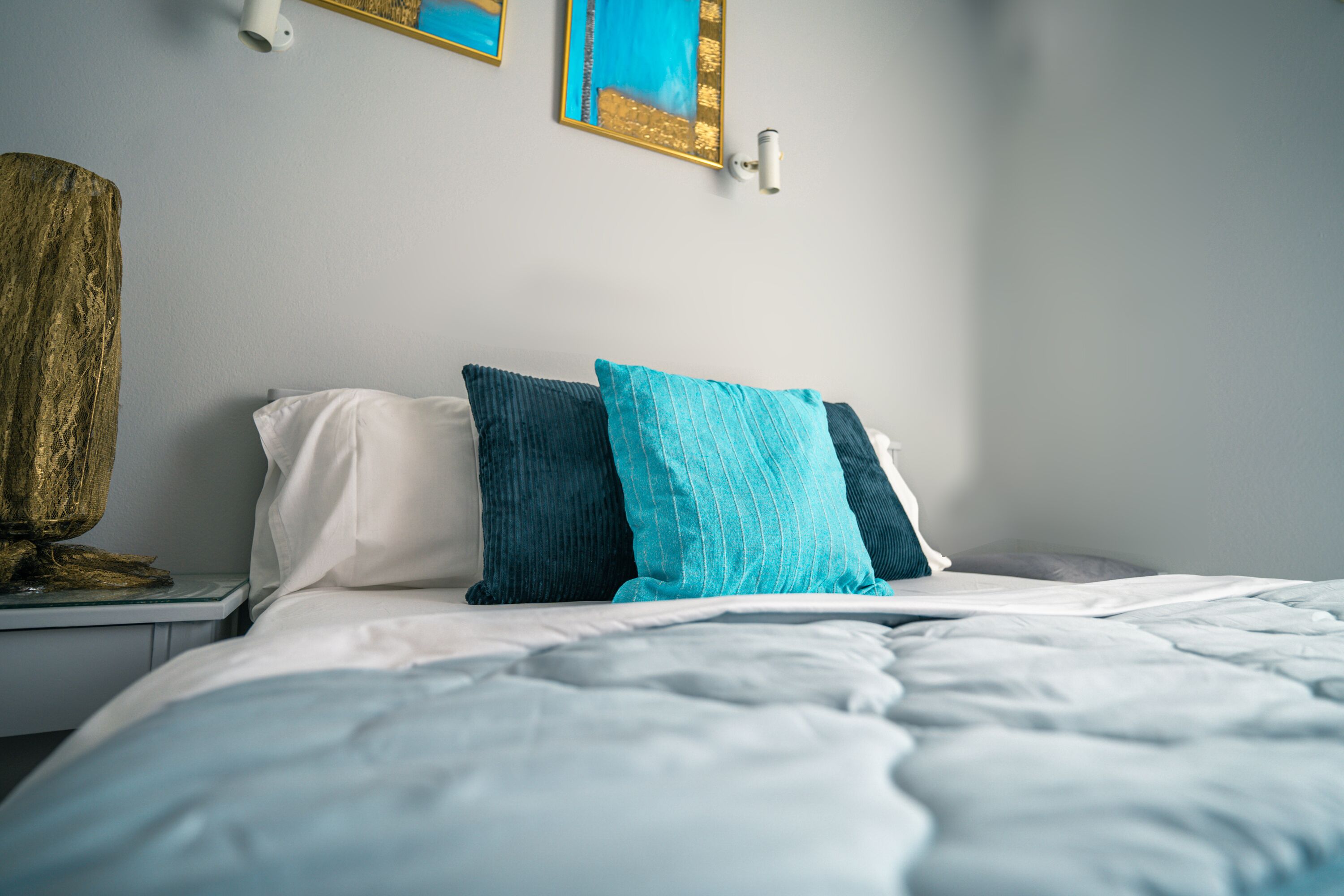 A neatly made bed with white and teal pillows, a wooden sculpture on the nightstand, and a framed picture on the wall.
