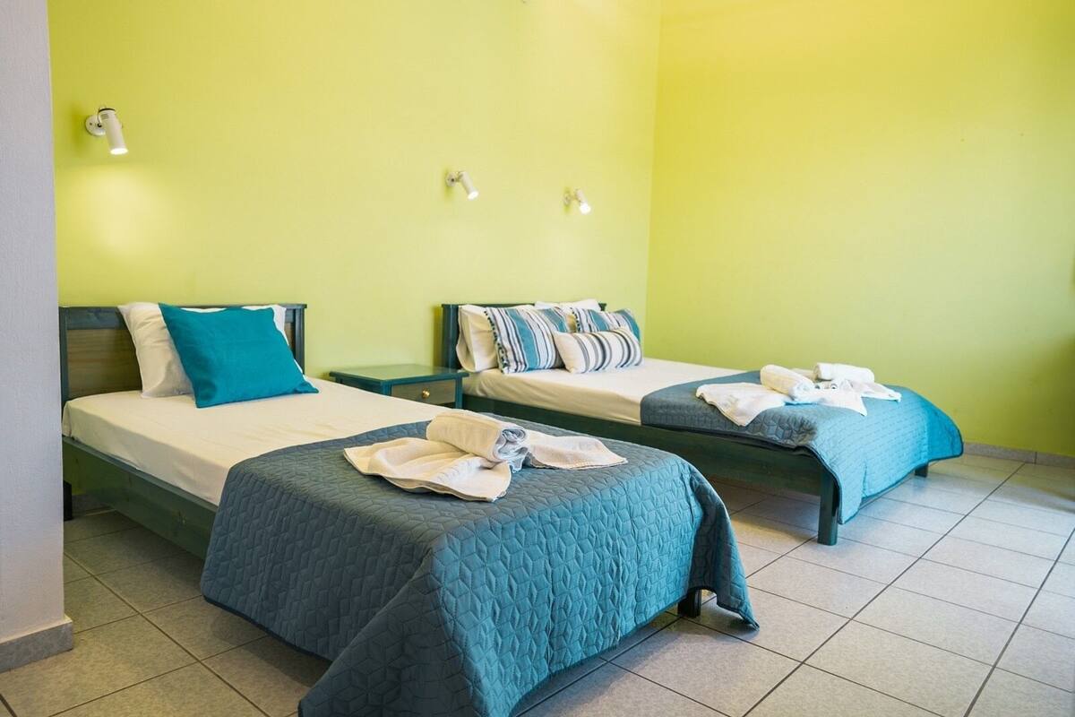 Quarto de hotel com duas camas, cada uma com colcha azul acolchoada e almofadas, uma pequena mesa de cabeceira e um candeeiro fixado na parede.