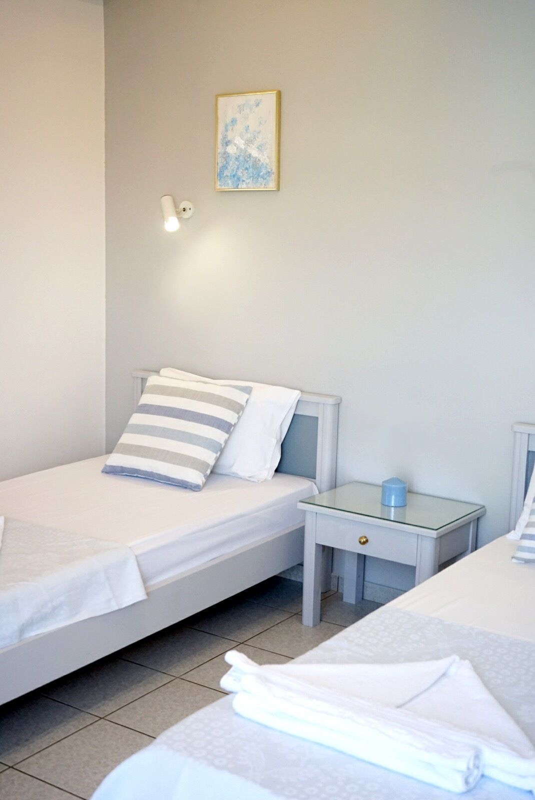 A hotel room with two single beds, a small bedside table with a lamp, and a framed picture on the wall.