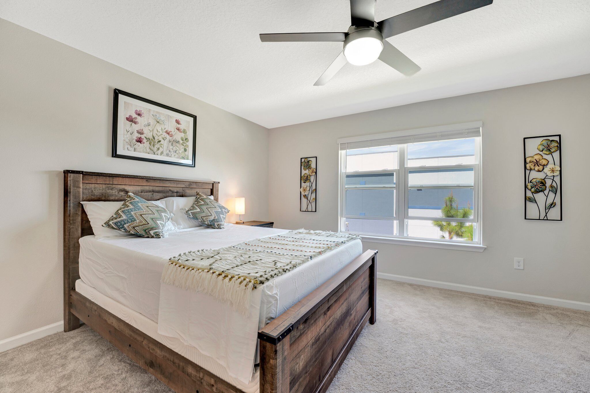 A bedroom with a bed, a ceiling fan, a window with a view, and floral artwork on the walls.