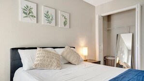 Habitación de hotel con cama, ropa de cama blanca, manta azul y tres impresiones botánicas enmarcadas en la pared.