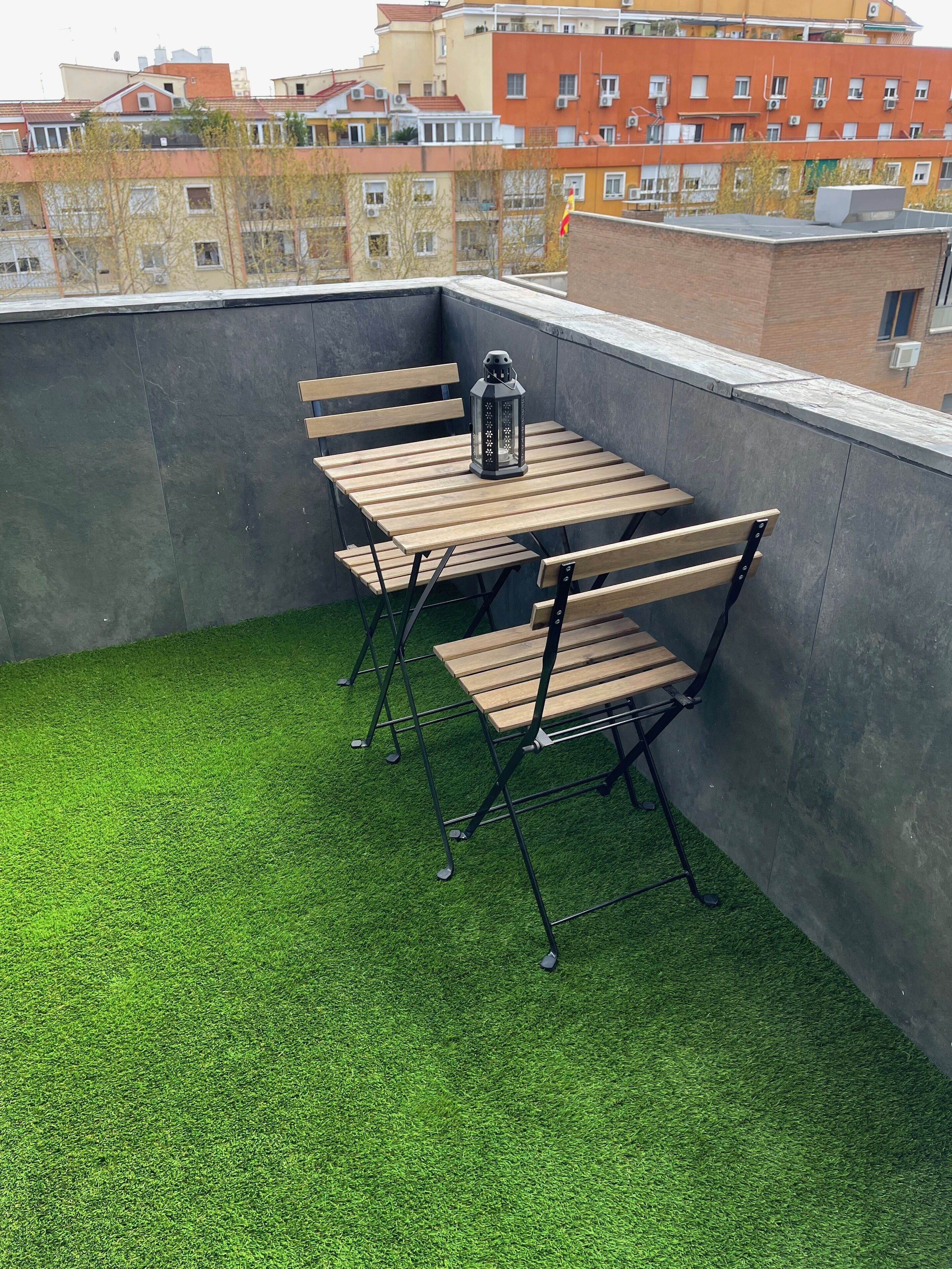 A rooftop terrace with a wooden table and chairs, a lantern, and artificial grass.