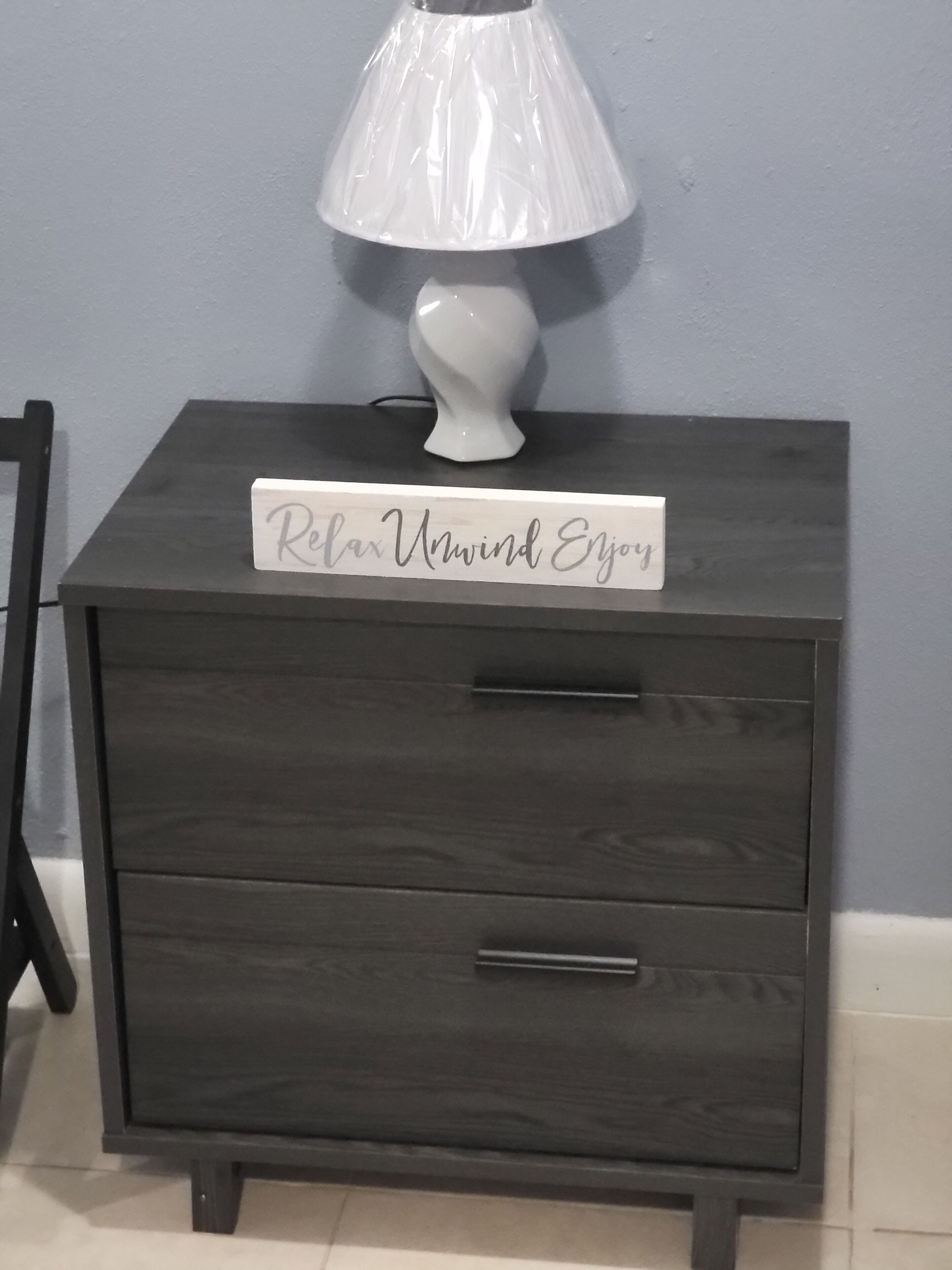 Un comodino in legno scuro con una lampada bianca e un cartello con su scritto 'Relax Unwind Enjoy'.