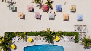 Vue aérienne d’un complexe hôtelier en bord de mer, avec une piscine, des parasols de plage, des chaises longues et des palmiers.