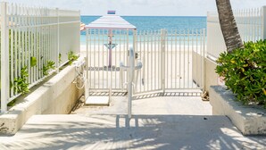 Un sentier bordé de barrières mène à une zone de plage agrémentée d’un palmier et d’un kiosque.