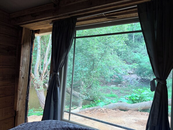 Quarto com uma grande janela, paredes de madeira e cortinas amarradas, revelando uma vista de uma área arborizada.