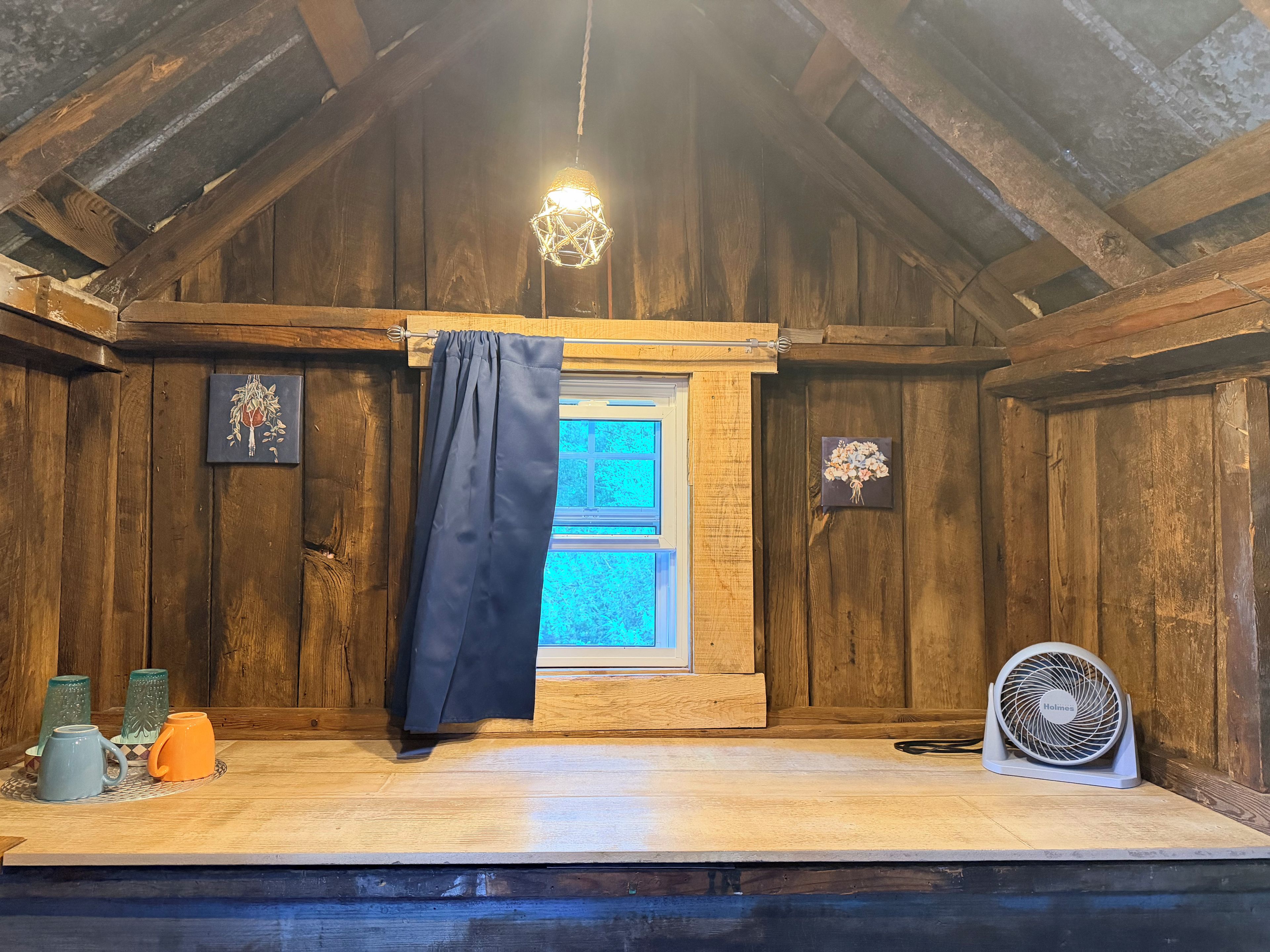 A rustic room with wooden walls, a small window, a hanging light fixture, and a fan on the floor.