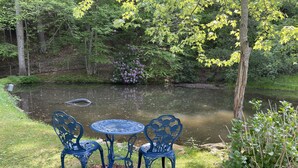 A serene outdoor seating area with a table and chairs by a pond, surrounded by lush greenery and trees. - Cozy Cabin: Peaceful Haven in the Heart of Nature (Green Mountain)