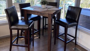 Comedor con mesa de madera y cuatro sillas, ventanas amplias con vista a un bosque y una pequeña planta en maceta sobre la mesa.