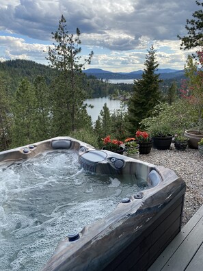 Un bain à remous entouré d’une terrasse agrémentée de plantes en pot, avec vue sur un lac et des montagnes boisées.