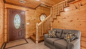 Un interior de madera con una escalera, un sofá gris con cojines estampados y una puerta con un vitral.