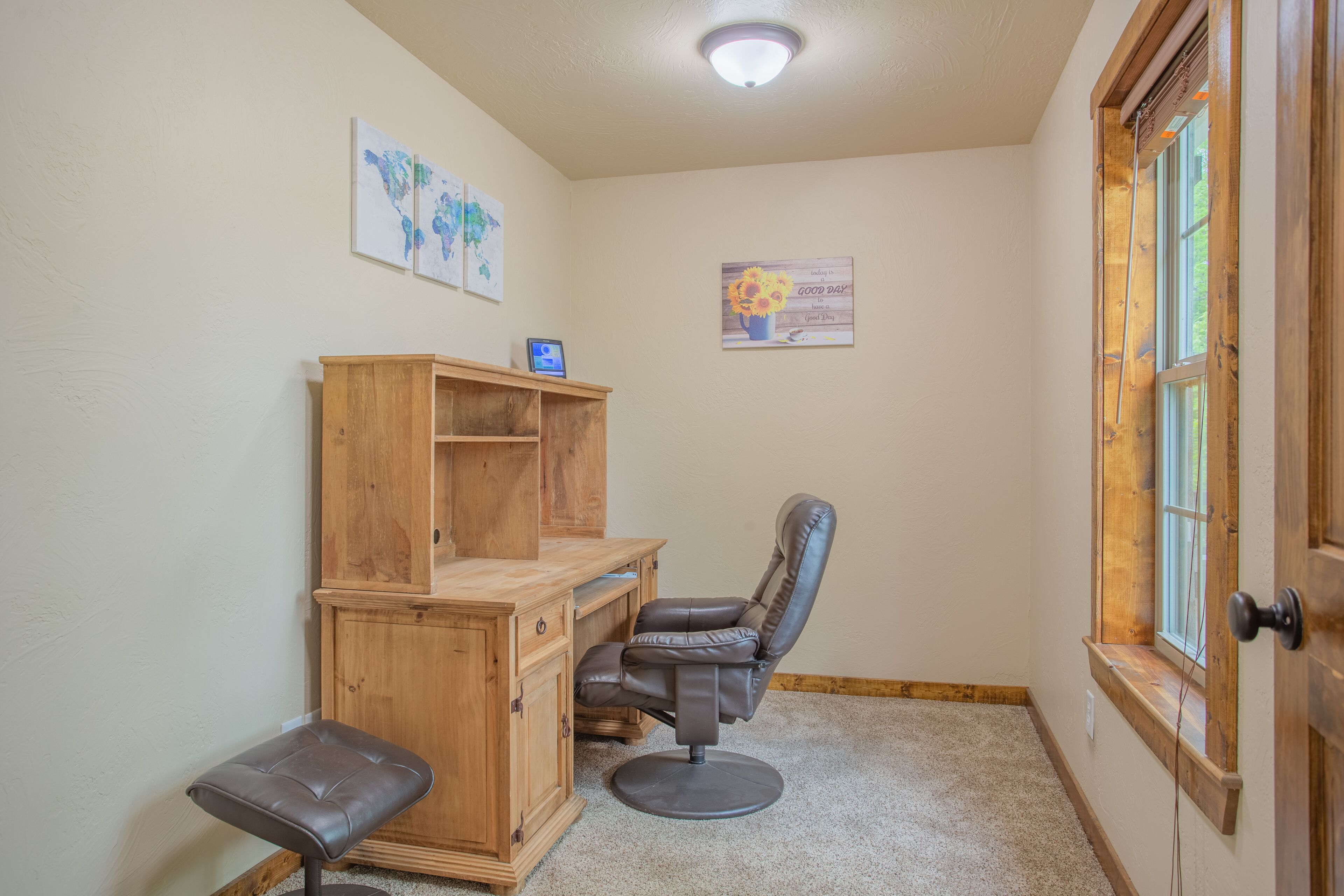 A corner room with a wooden desk, a leather chair, a small stool, and a window with a wooden frame.