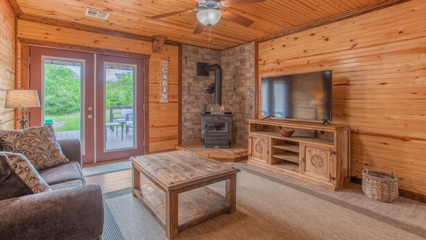 Interior de una cabaña con techo de madera, ventilador de techo, chimenea de piedra, televisor, mesa de madera y sofá.