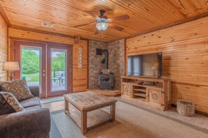 A cabin interior with a wooden ceiling, a ceiling fan, a stone fireplace, a television, a wooden table, and a sofa.