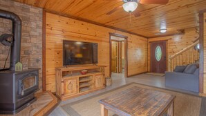 Interior de una cabaña de troncos con chimenea, televisor, sofá y escalera.