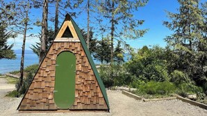 A triangular wooden structure with a green door, surrounded by trees and a clear sky. - A-frame Shelter (T16) Oceanview Olympic Peninsula (Port Angeles)