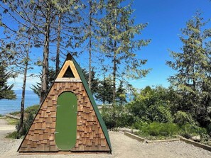 A triangular wooden structure with a green door, surrounded by trees and a clear sky. - A-frame Shelter (T16) Oceanview Olympic Peninsula (Port Angeles)