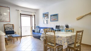 Un comedor con una mesa dispuesta para cuatro, un sofá azul, una silla de madera con un cojín estampado y un balcón con vistas.